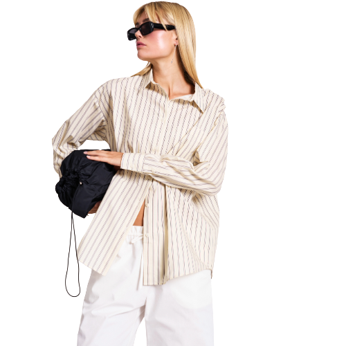 Woman wearing sunglasses, an oversized beige striped shirt, and white pants holds a black bag. She stands against a plain white background, looking off to the side with a casual pose.
