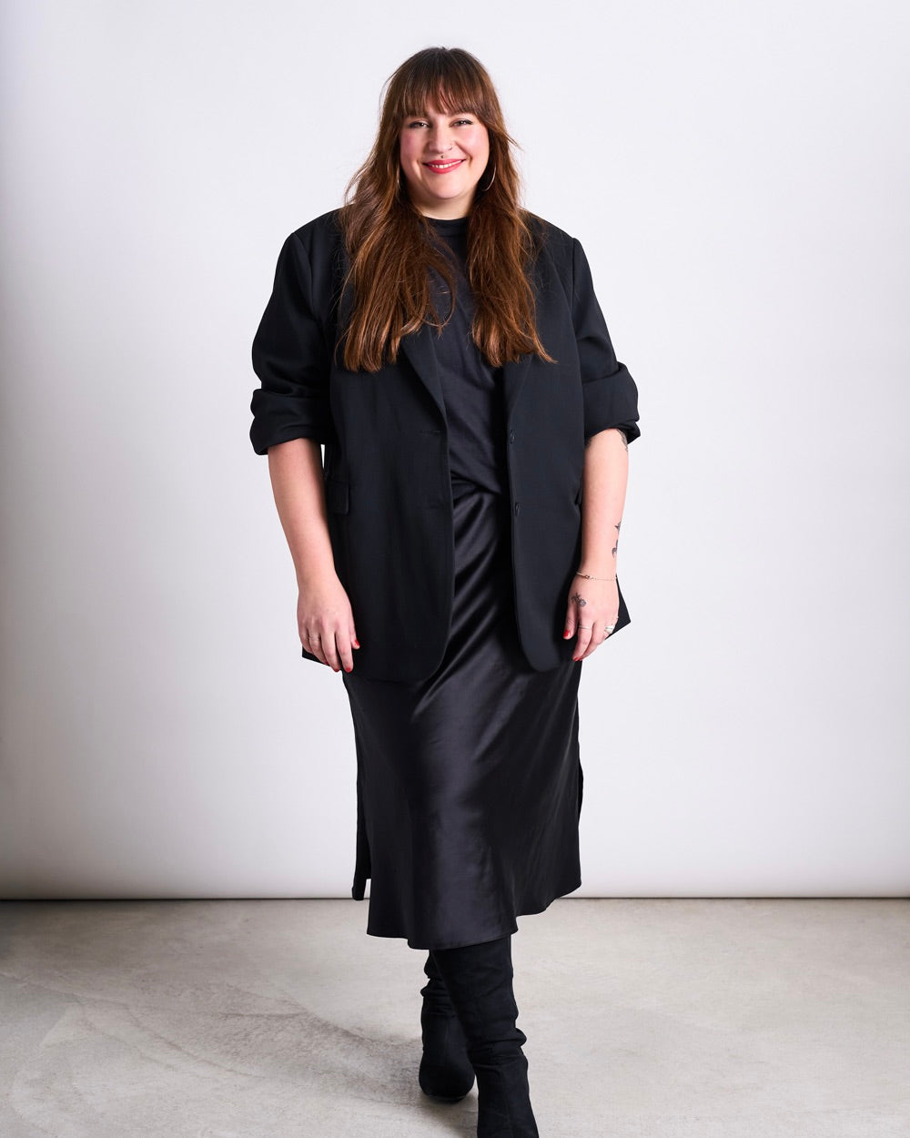 Woman standing in front of a light background wearing a long black blazer, a black mid‑calf dress and black boots, looking at the camera and smiling.