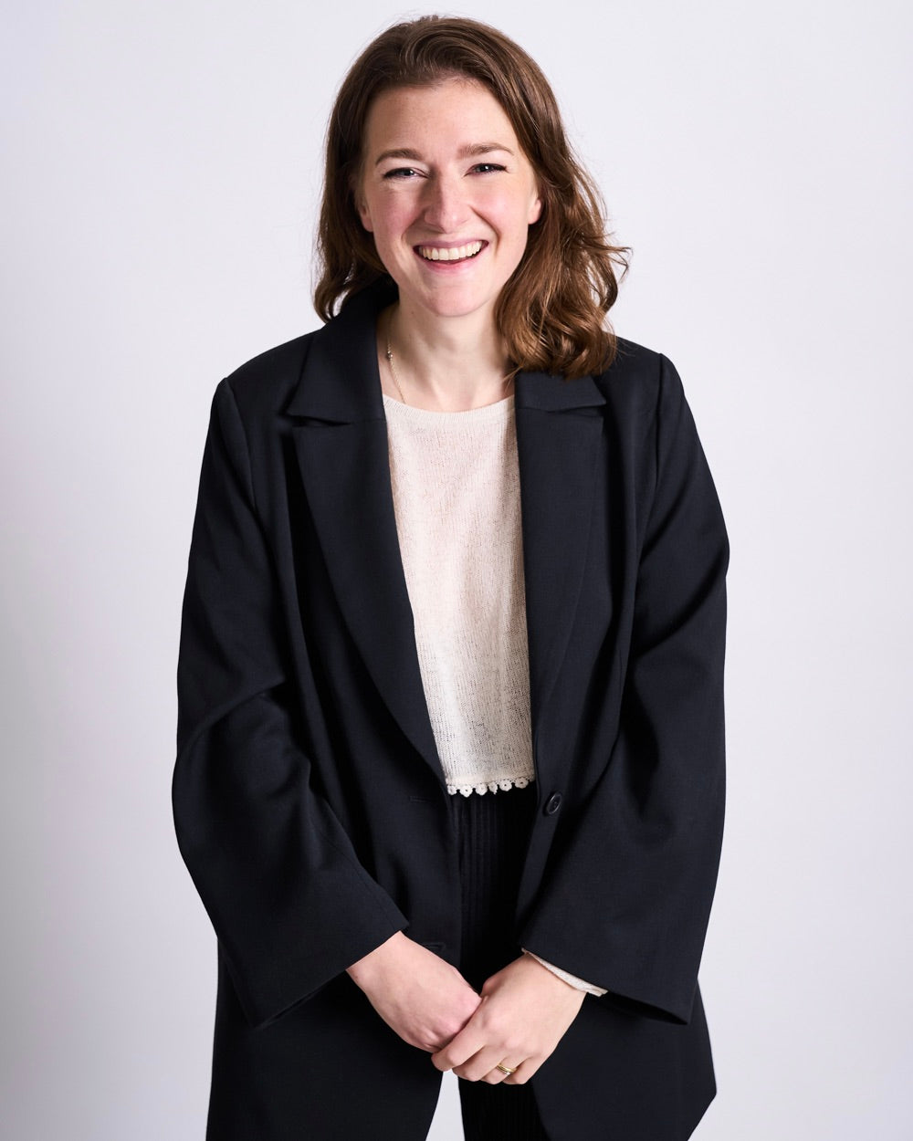 Woman standing against a light background wearing a black blazer over a light blouse and dark trousers, shoulder‑length brown hair, hands clasped in front.