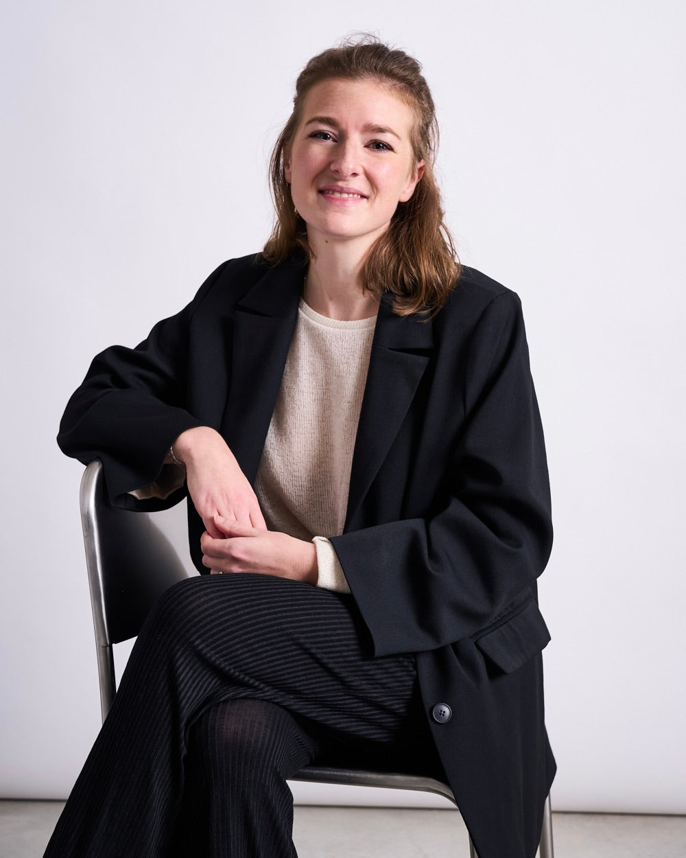 Woman seated on a chair positioned sideways, wearing a light beige sweater under a dark blazer and dark plain trousers, hands resting in their lap.