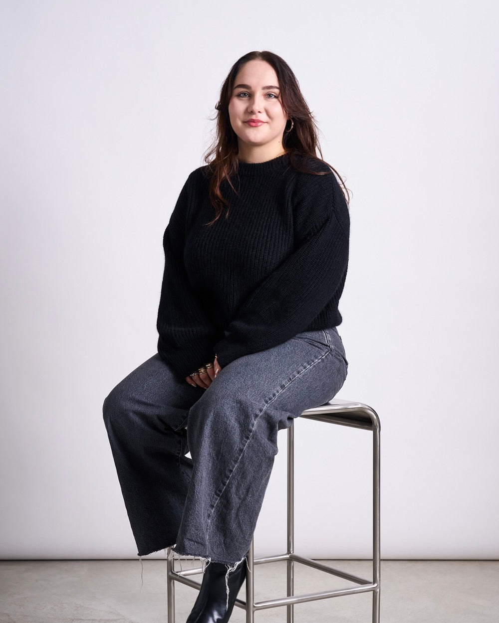 Woman seated on a tall metal stool wearing a black sweater, grey frayed‑hem jeans and black boots.