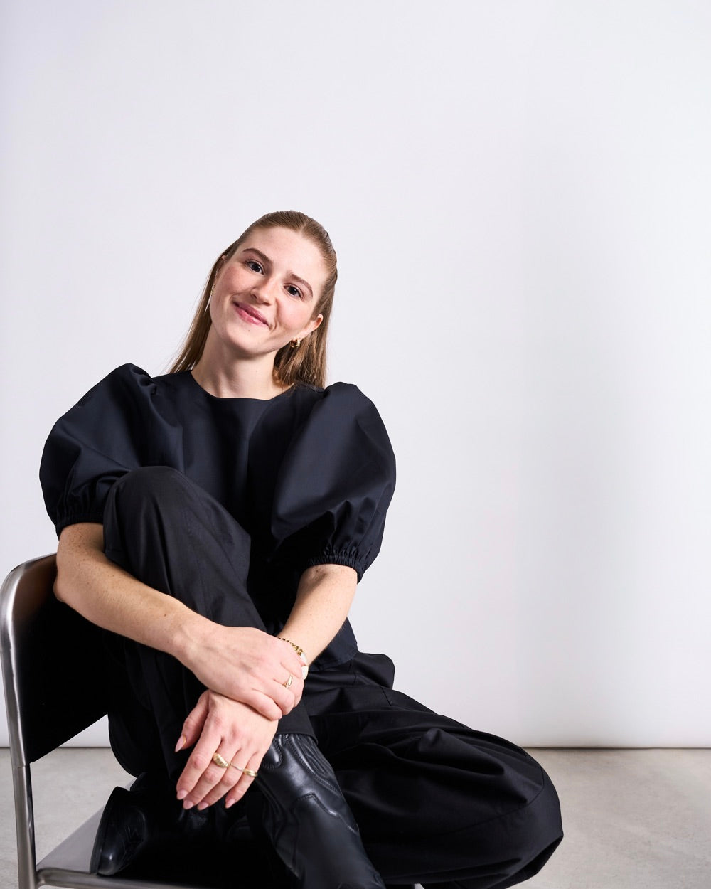 Woman seated on a chair wearing a black puff‑sleeve blouse and loose black trousers.