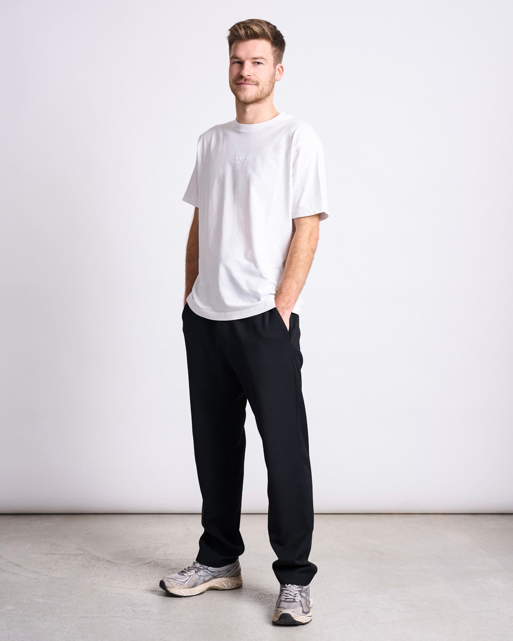 Man standing against a light grey background wearing a white T‑shirt, black trousers and light grey sneakers, both hands in their pockets.