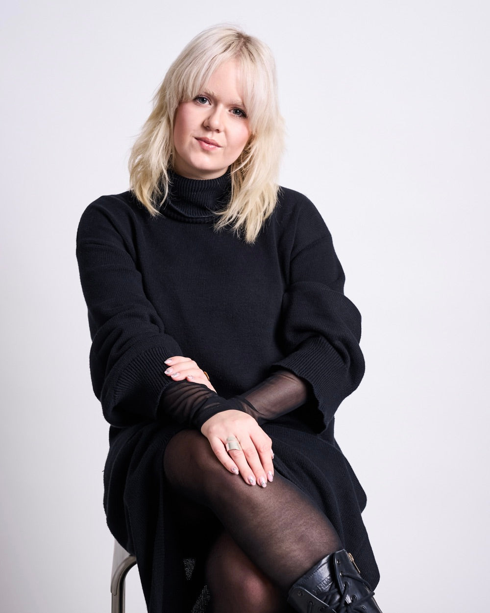 Woman seated on a stool wearing a long black turtleneck sweater dress, sheer black tights and black boots, light blonde hair with bangs, hands resting on the knee with a ring visible, against a plain light grey background.