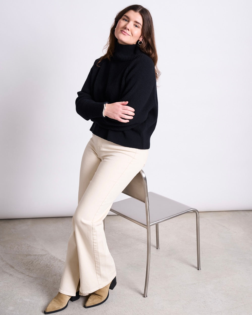 Woman sitting on the edge of a metal chair wearing a black turtleneck, cream wide trousers and tan boots.