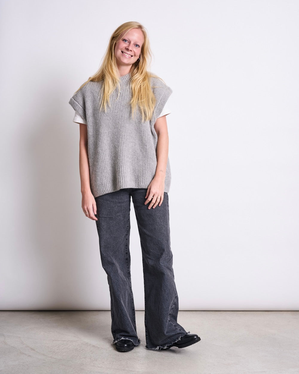Woman standing against a white wall wearing a grey sleeveless knit over a white T‑shirt, dark grey wide‑leg jeans and black shoes.
