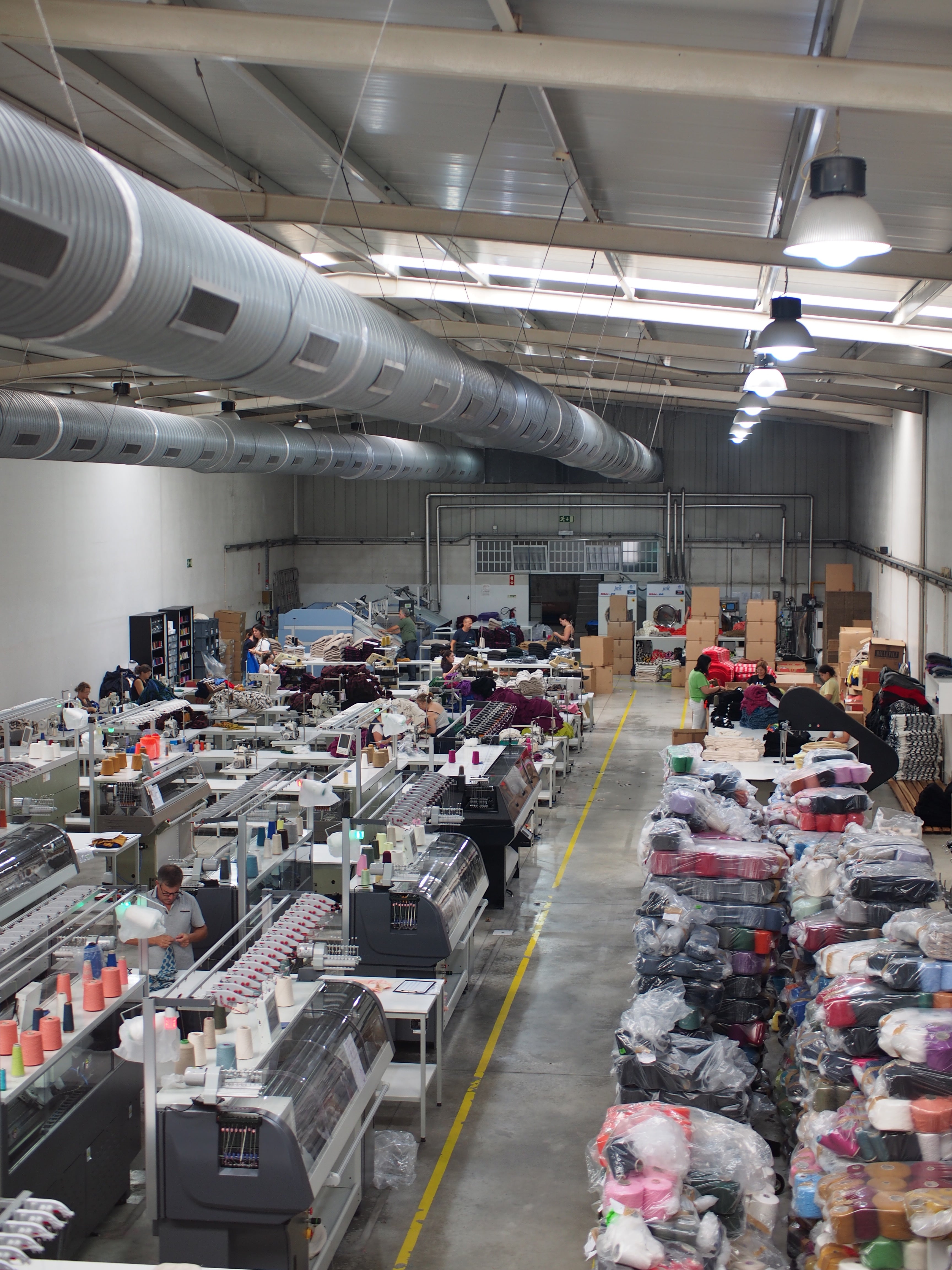 Interior of a textile production facility with rows of large weaving machines on one side, workers operating them, and stacks of filled bags and boxes on the other side, all in a brightly lit industrial space.