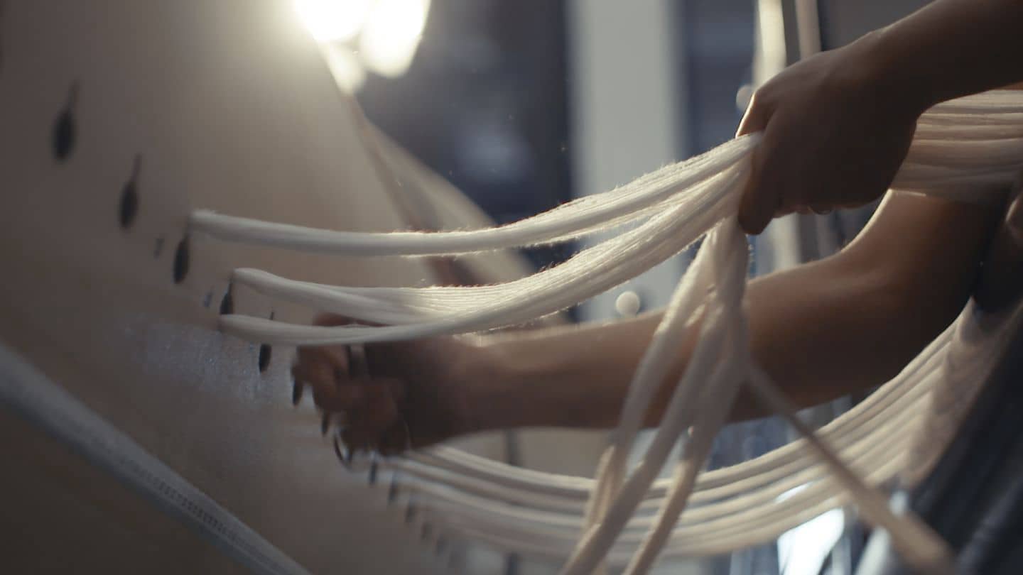 Close‑up of hands threading multiple white cords through a panel with evenly spaced holes, the strands soft and flexible, lit warmly.